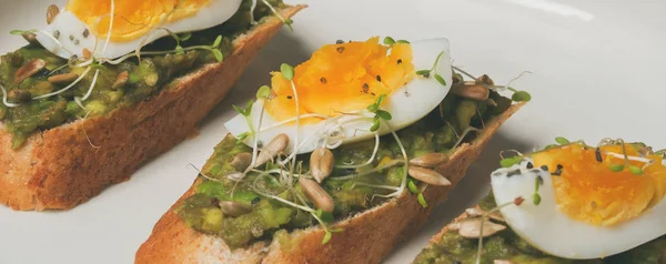 Whole-grain toasts plated avocado with eggs and fresh mini herbs with sunflower seeds and sprinkled black salt served on ceramic white plate over grey concrete background, top view, Diet food concept, gluten-free