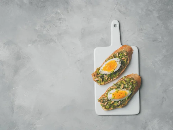 Whole-grain toasts plated avocado with eggs and fresh mini herbs served on ceramic white board over grey concrete background, top view, Diet food concept, gluten-free