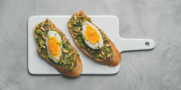 Top view of whole-grain toasts plated avocado with eggs, fresh mini herbs, sunflower seeds and sprinkled black salt served on ceramic white board