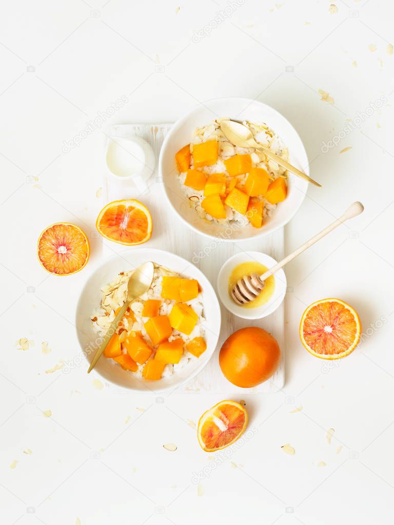 rice porridge bowls with coconut milk served with mango and almond petals, honey and oranges on white background