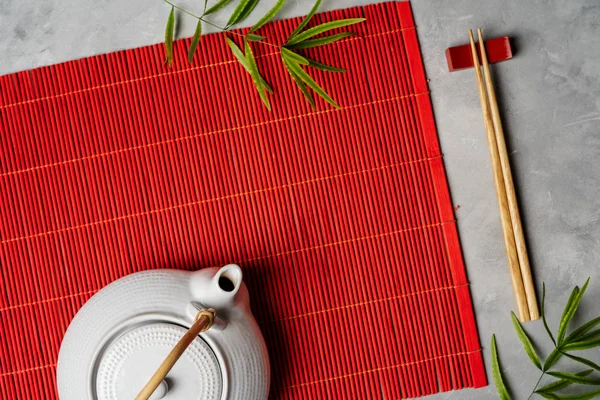 Asian tea set and chopsticks on red bamboo mat on gray stone background