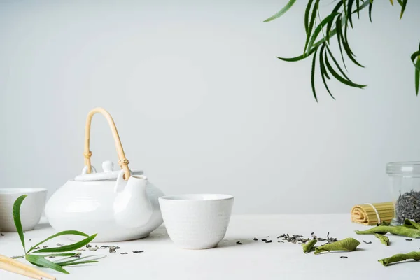 ライトグレーの背景に葉の茶と緑茶乾燥で囲まれたティーポットの つの白いカップでアジアのお茶セット — ストック写真