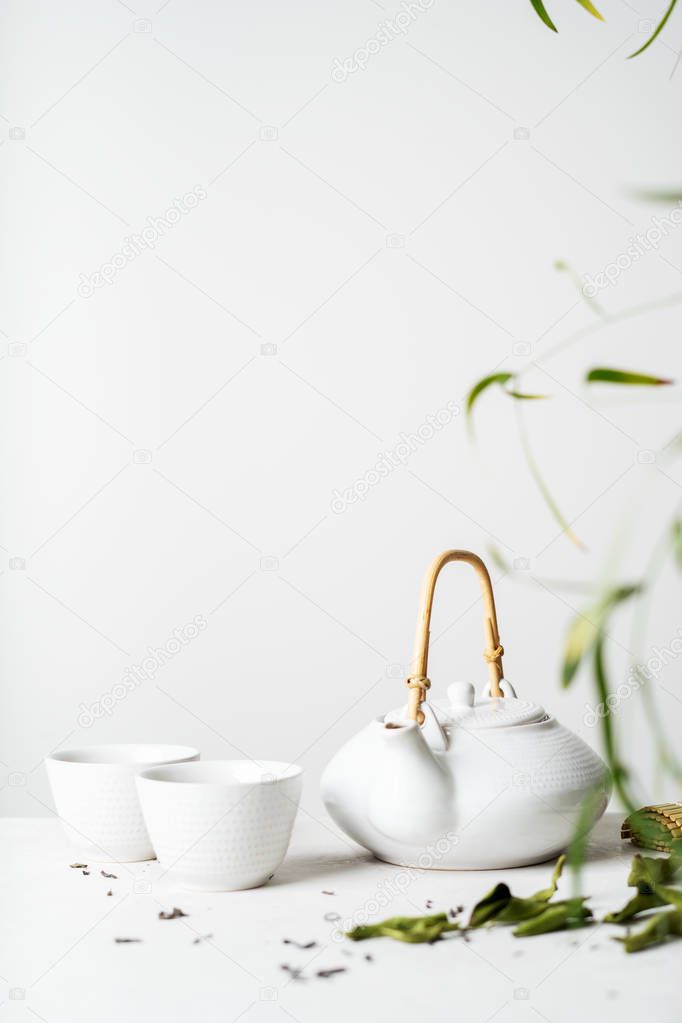 Asian tea set with two white cups of tea and teapot surrounded with green tea dry leaves on light grey background