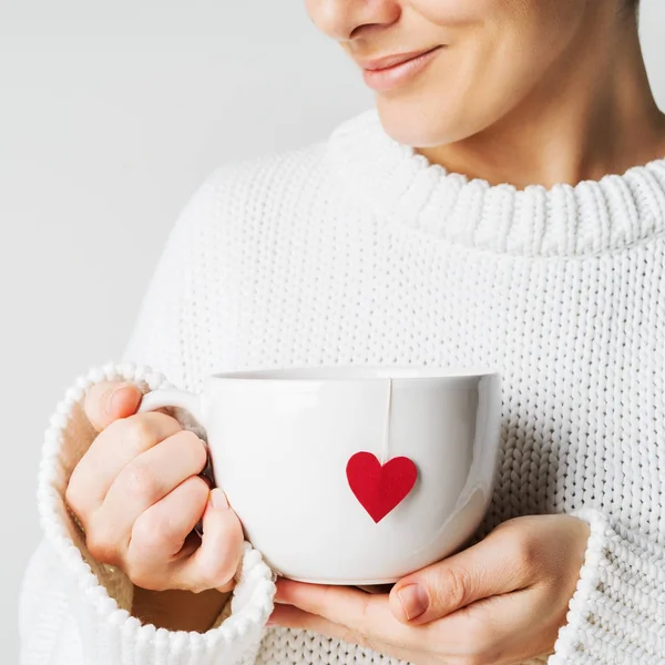 Close view of woman in warm knitted woolen sweater holding white mug in hands with heart of tea bag with love, Valentine\'s day mockup
