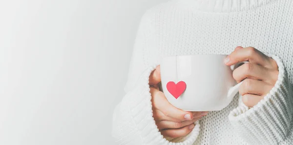 Close view of woman in warm knitted woolen sweater holding white mug in hands with heart of tea bag with love, Valentine\'s day mockup