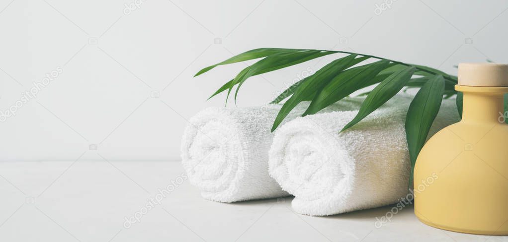two white fluffy towels twisted into rolls and ceramic bottle on light surface with palm leaf