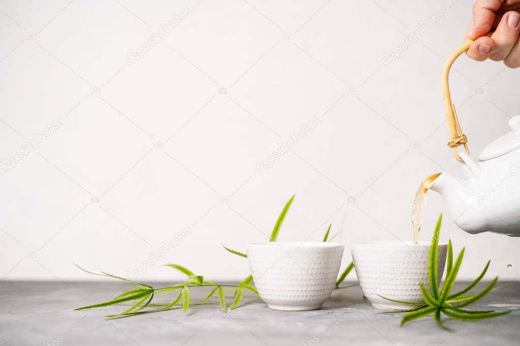Female hand pouring green tea from a teapot into cups on white background with copy space. Asian tea set. 