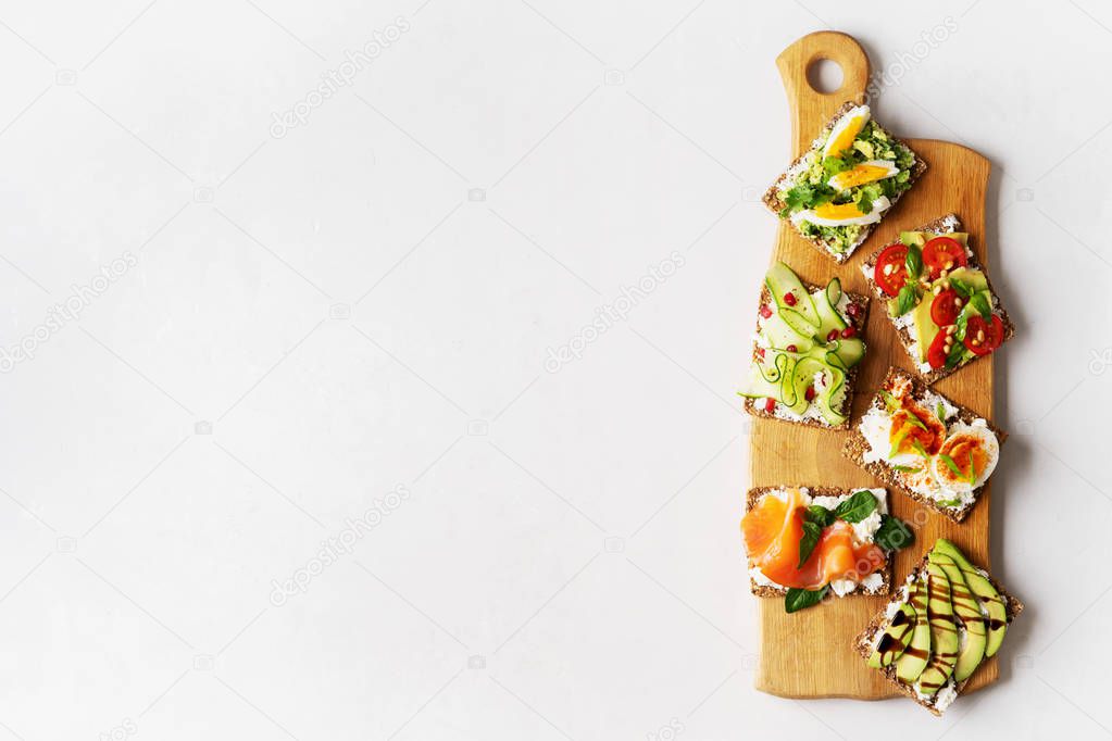 Top view of protein toasts with smoked salmon, spinach, cucumber, tomatoes and cream cheese on wooden long cutting board over light grey background