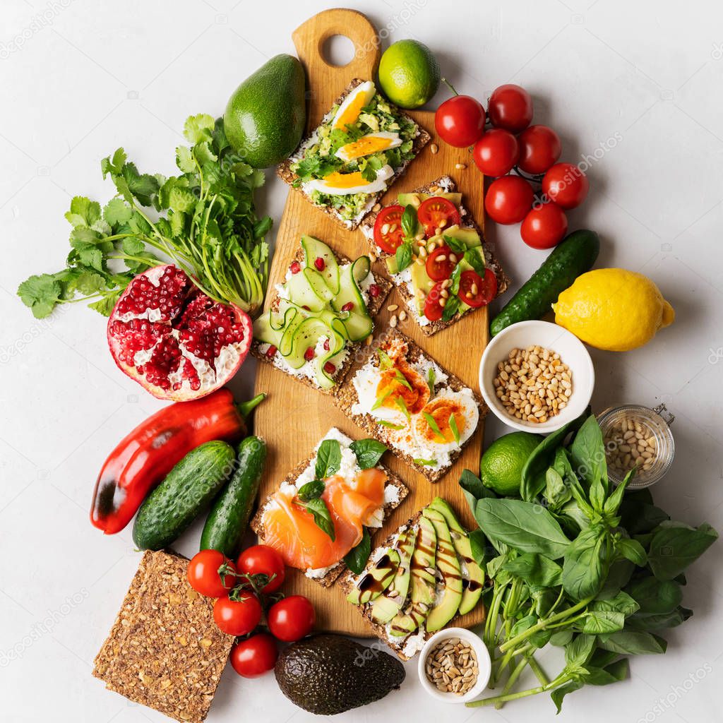Top view of Superfood protein toast with delicious toppings on top: smoked salmon, spinach, cucumber, tomato and cream cheese on wooden long cutting board among vegetables ingredients. healthy snacks.