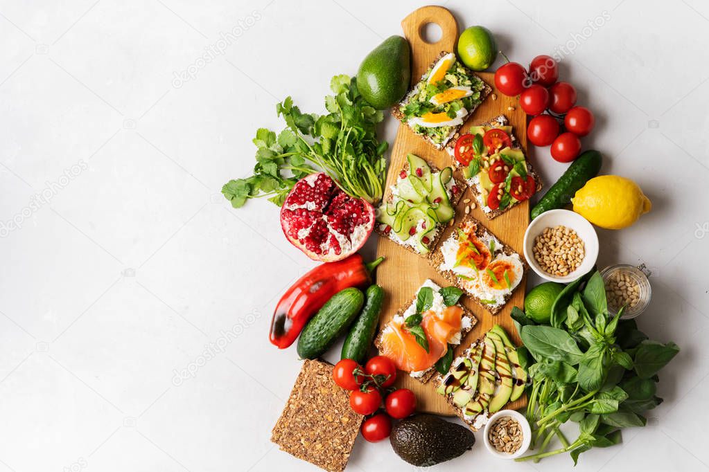Top view of Superfood protein toast with delicious toppings on top: smoked salmon, spinach, cucumber, tomato and cream cheese on wooden long cutting board among vegetables ingredients. healthy snacks.