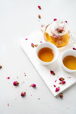 iki beyaz bardak ve gri taş arka planda kurutulmuş pembe gül tomurcukları bitkisel çay ile şeffaf çaydanlık