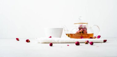 Beyaz seramik fincan ve gri arka planda kurutulmuş pembe güller bitkisel çay ile şeffaf çaydanlık