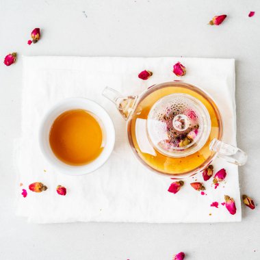 Beyaz seramik fincan ve gri arka planda kurutulmuş pembe güller bitkisel çay ile şeffaf çaydanlık