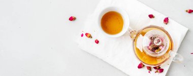 Beyaz seramik fincan ve gri arka planda kurutulmuş pembe güller bitkisel çay ile şeffaf çaydanlık