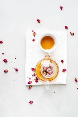 Beyaz seramik fincan ve gri arka planda kurutulmuş pembe güller bitkisel çay ile şeffaf çaydanlık