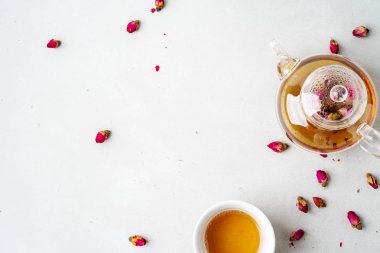 Beyaz seramik fincan ve gri arka planda kurutulmuş pembe güller bitkisel çay ile şeffaf çaydanlık