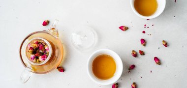 iki beyaz bardak ve gri taş arka planda kurutulmuş pembe gül tomurcukları bitkisel çay ile şeffaf çaydanlık