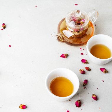 iki beyaz bardak ve gri taş arka planda kurutulmuş pembe gül tomurcukları bitkisel çay ile şeffaf çaydanlık