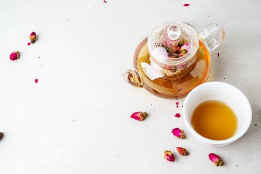 Beyaz seramik fincan ve gri arka planda kurutulmuş pembe güller bitkisel çay ile şeffaf çaydanlık