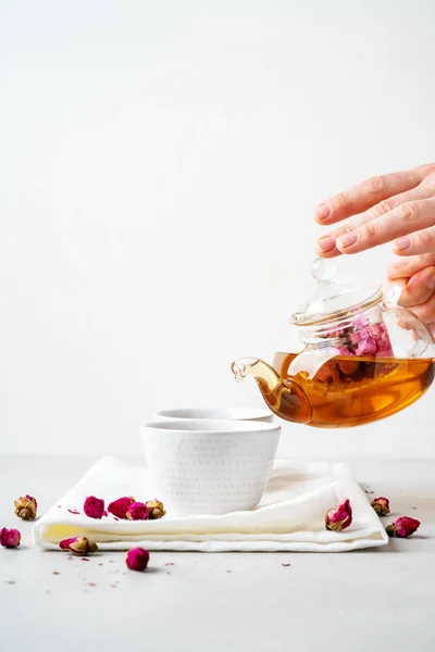 Female hands pouring tea of dry pink rose buds from transparent kettle to teacups on white background