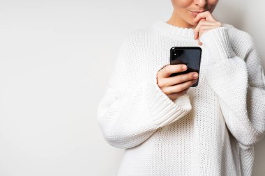 Beyaz arka planda akıllı telefonu elinde tutan beyaz kazaklı genç kadının yakın görünümü 