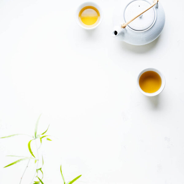 Two white cups of tea and teapot surrounded with green leaves on white background