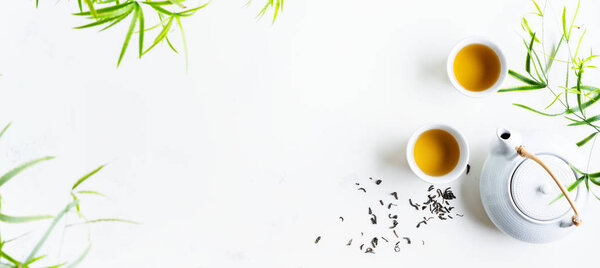 Two white cups of tea and teapot surrounded with green leaves on white background