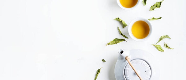 Two white cups of tea and teapot surrounded with green leaves on white background