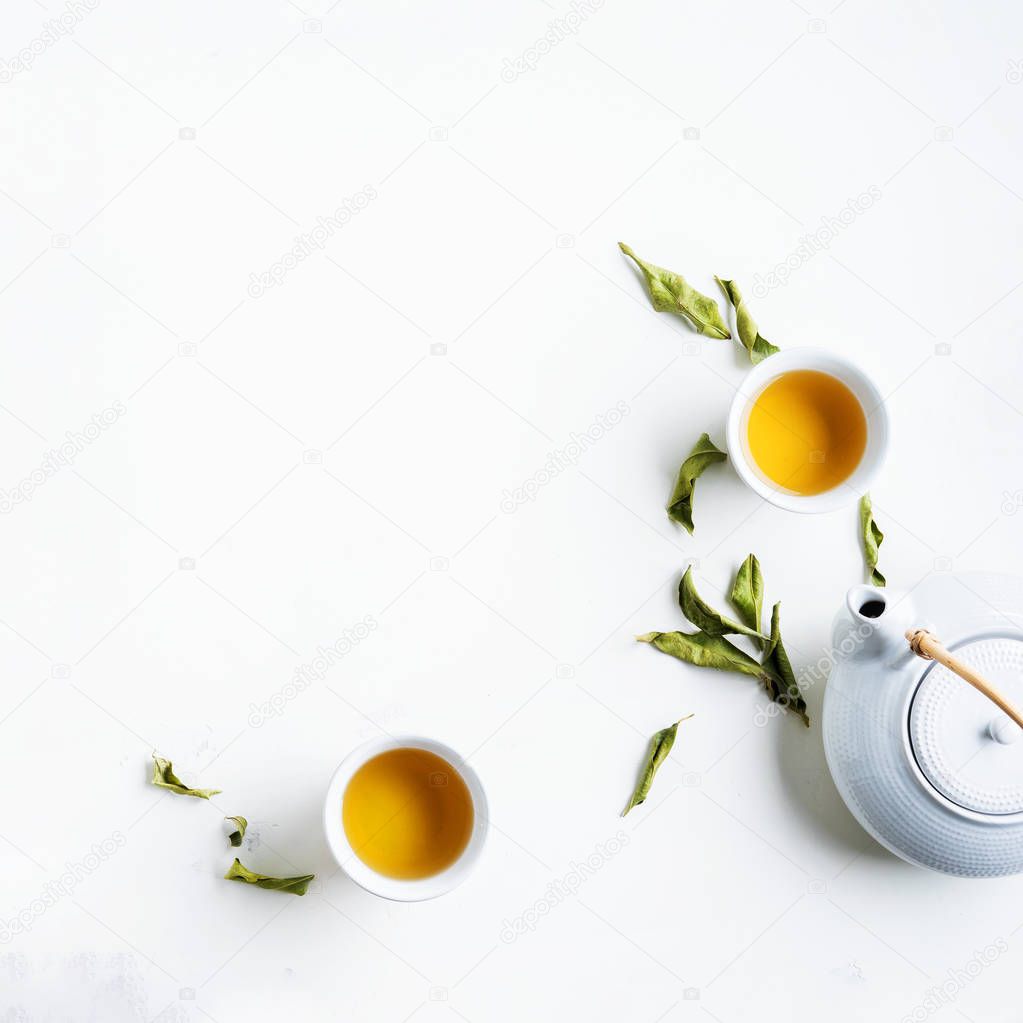 Two white cups of tea and teapot surrounded with green leaves on white background