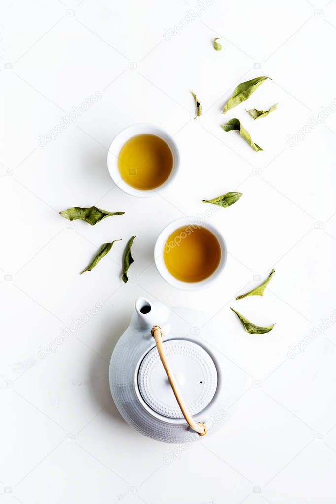 Two white cups of tea and teapot surrounded with green leaves on white background