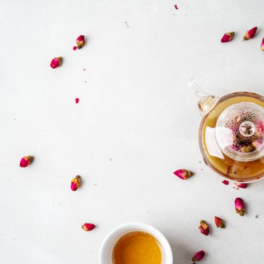 Beyaz seramik fincan ve gri arka planda kurutulmuş pembe güller bitkisel çay ile şeffaf çaydanlık