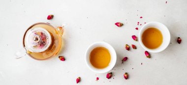 iki beyaz bardak ve gri taş arka planda kurutulmuş pembe gül tomurcukları bitkisel çay ile şeffaf çaydanlık