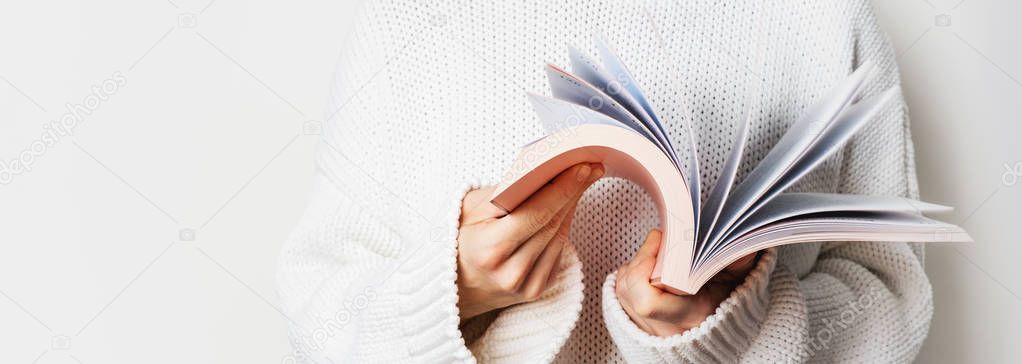 Close view of woman in white woolen sweater holding open book with pink cover on light background 