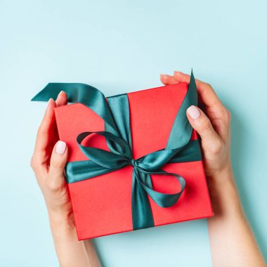 Pastel mavi arka plan üzerinde yeşil yay ile mevcut kırmızı kutu tutan kadın eller