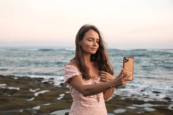 Beautiful girl preens looking at the phone on the beach on a background of the sea. A nice young girl straightens her hair while looking in a cell phone.