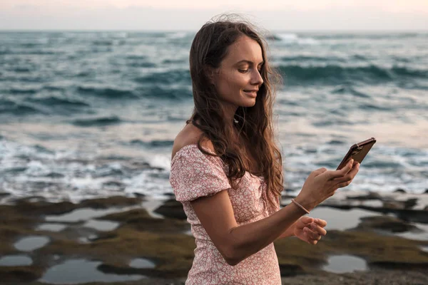 Beautiful girl preens looking at the phone on the beach on a background of the sea. A nice young girl straightens her hair while looking in a cell phone.