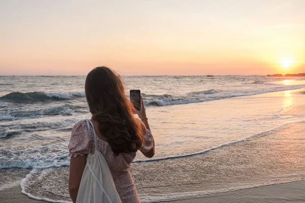 Gün batımında kadın eli cep telefonu tutuyor. Deniz aşırı güzel bir gün batımında cep telefonuyla fotoğraf çeken genç bir kadın. Endonezya, Bali 'de bir tatil sırasında akıllı telefondan fotoğraf çekmek