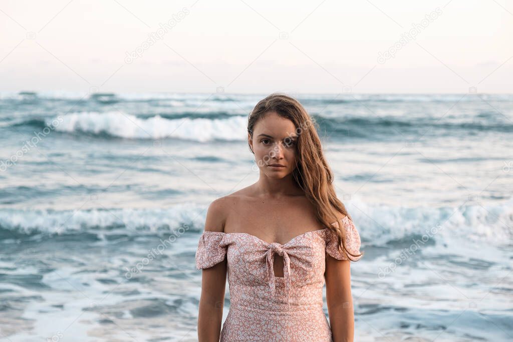 A sad beautiful young girl in a dress stands on the ocean and looks at the camera. Pleasant pastel colors. Girl close-up on a background of waves. The woman turned her back to the sea