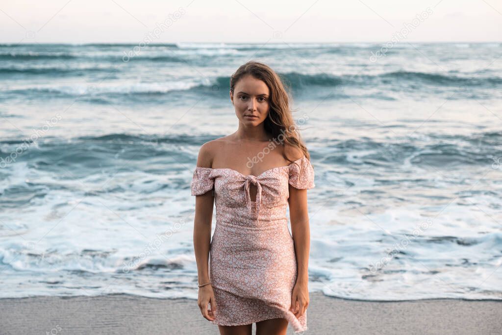 A sad beautiful young girl in a dress stands on the ocean and looks at the camera. Pleasant pastel colors. Girl on a background of waves. The woman turned her back to the sea
