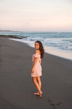 Genç ve güzel bir turist kız Bali 'de okyanus kıyısında boş bir kumsal boyunca yürüyor. Elbiseli kız gülümser ve arkasını döner..