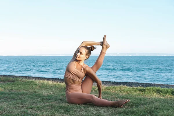 Genç ve güzel bir kadın doğada yoga yapıyor ve sicimde oturuyor, bir bacağını başının üstüne kaldırıyor. Yoga hocası, okyanus kenarındaki çimlerde yoga yapıyor..