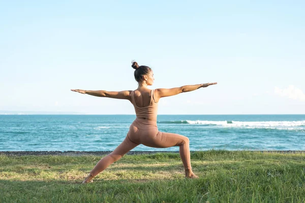 Young beautiful woman is practicing warrior pose yoga, standing in Warrior Two exercise, Virabhadrasana pose. Girl doing yoga on the grass by the sea