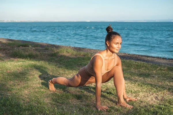 Spor giyim sektöründe bir kadın yogi okyanusun arka planında çimlere vuruyor. Bir kadın tek ayağının üzerine atlıyor ve arkasına yaslanıyor. Kız okyanusta yoga yapıyor. Sağlık ve uyum için spor ve esneme