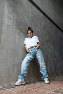 Büyüme çağında güzel bir kadın duvarın önünde duruyor. Mavi kotlu ve beyaz tişörtlü şık bir kız gri beton bir duvarın arkasında duruyor. 80 'lerin tarzı