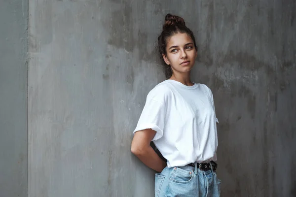 Üzgün düşünceli bir kız duvarda duruyor. Mavi kotlu ve beyaz tişörtlü şık bir kadın gri beton bir duvarın arkasında duruyor..