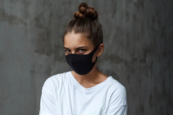 Siyah koruyucu maskeli güzel bir kadın kameraya bakıyor. Virüsten koruma maskesi takmış meraklı bir kadın gri beton bir duvarın arkasında duruyor.