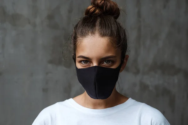 Siyah koruyucu maskeli güzel bir kadın kameraya bakıyor. Virüsten koruma maskesi takmış meraklı bir kadın gri beton bir duvarın arkasında duruyor.