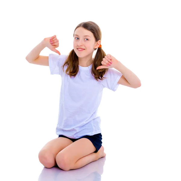Kleines Schulmädchen zeigt auf ihr leeres weißes T-Shirt. — Stockfoto