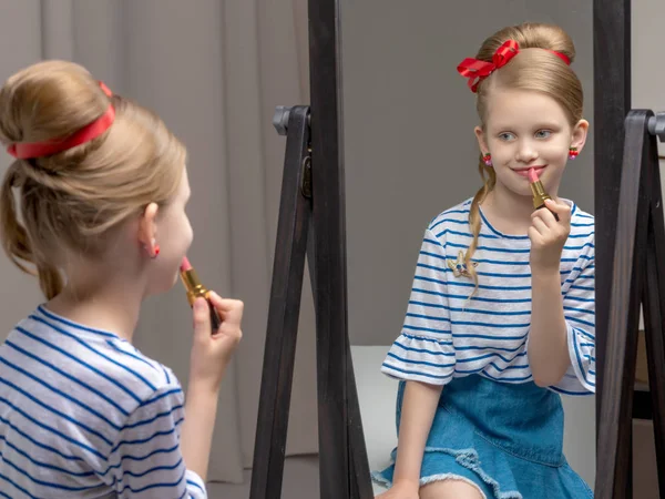 A little girl near the mirror paints lipstick lips — Stock Photo, Image