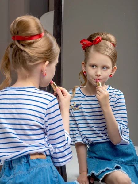 A little girl near the mirror paints lipstick lips — Stock Photo, Image